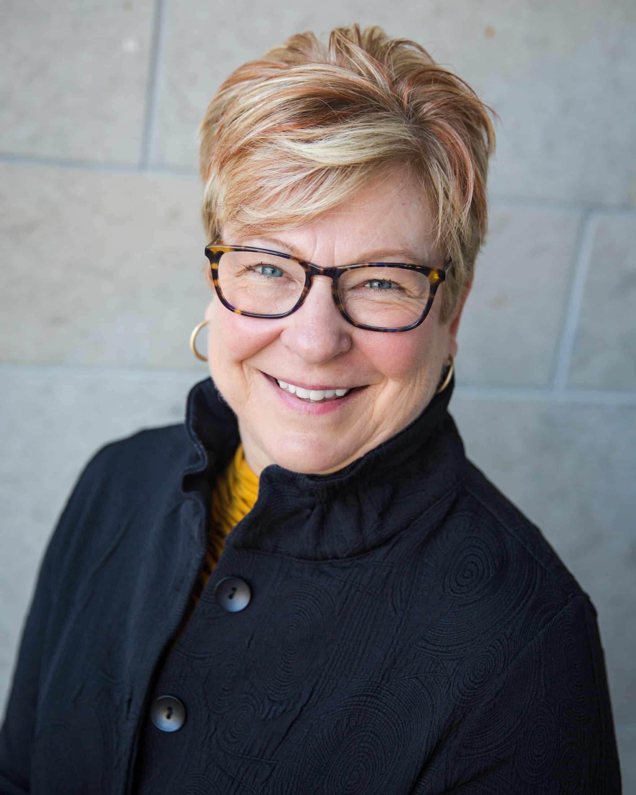 Woman smiling while wearing black button up jacket, round glasses, and hoop earrings